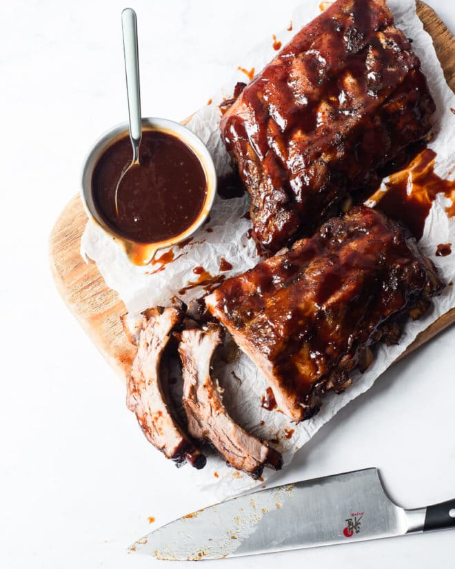 two half racks of ribs smothered in BBQ sauce sitting on a wooden cutting board next to a bowl of more BBQ sauce