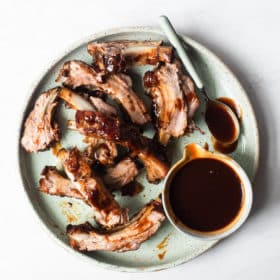 a light green plate of slow cooker baby back ribs (separated) next to a bowl of BBQ sauce with a spoon sticking out of it