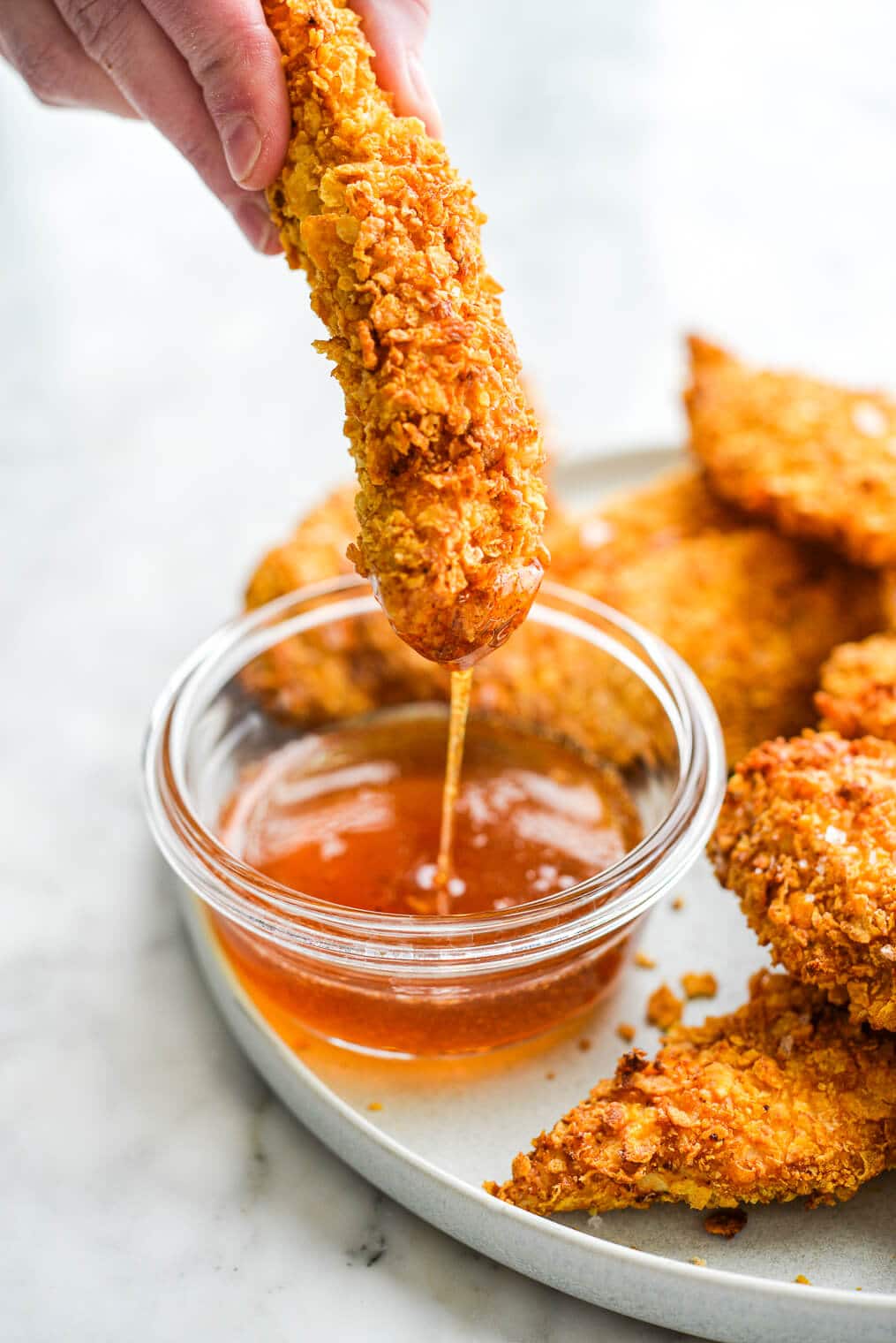 a person holding a crispy air fryer chicken tender above a small bowl of hot honey dipping sauce