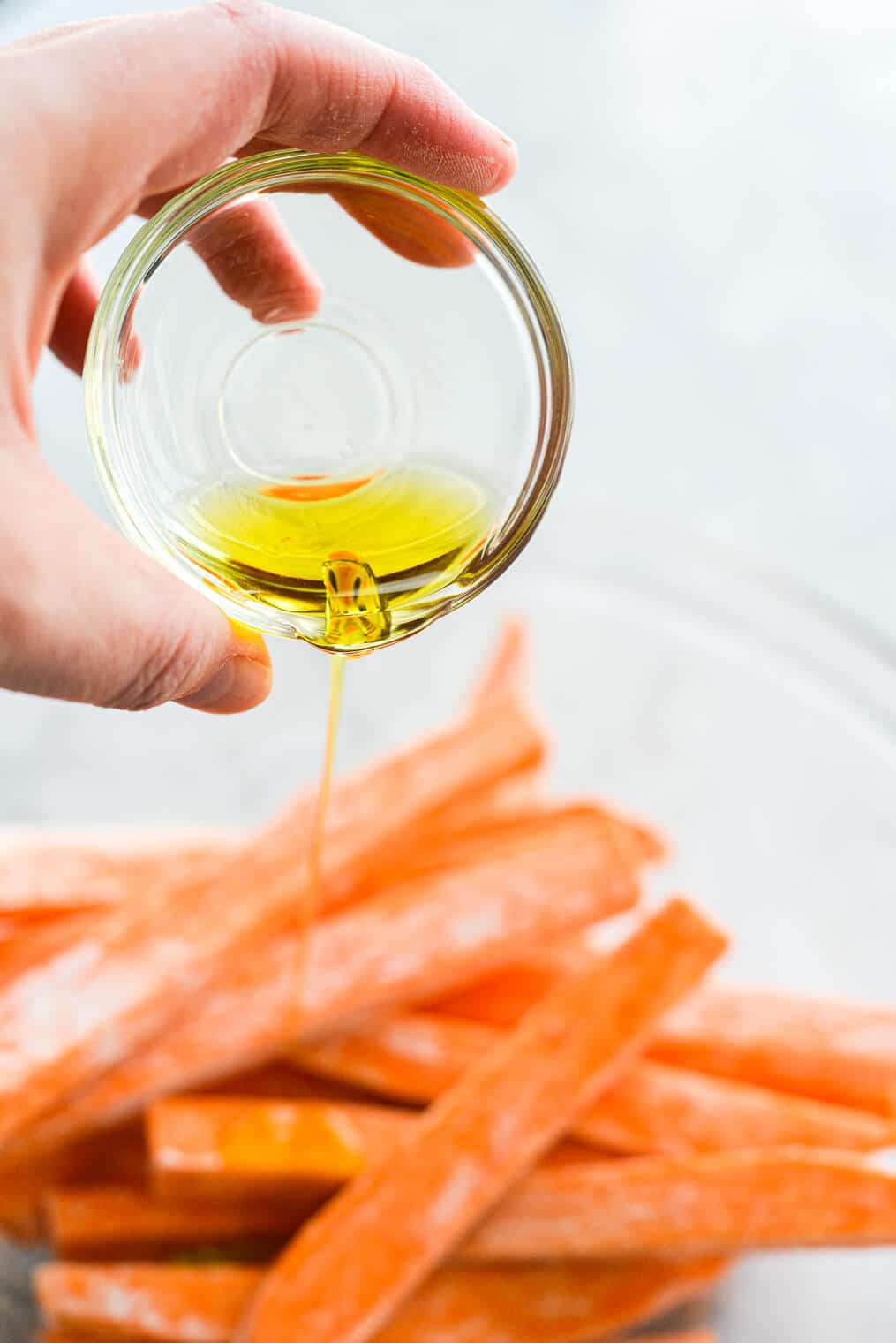 a person pouring a small clear bowl of olive oil over fry-cut sweet potatoes