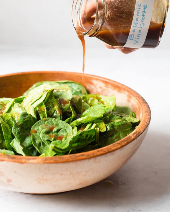 a homemade balsamic vinaigrette dressing being poured over a wooden bowl filled with spinach leaves