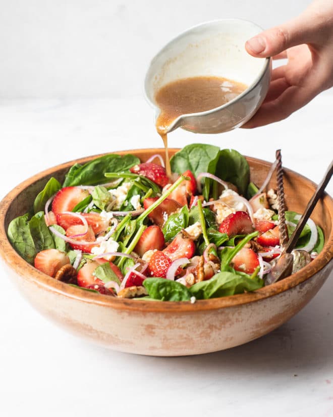 a large wooden bowl of strawberry spinach salad with two large spoons sticking out of it and a woman pouring dressing over top of it