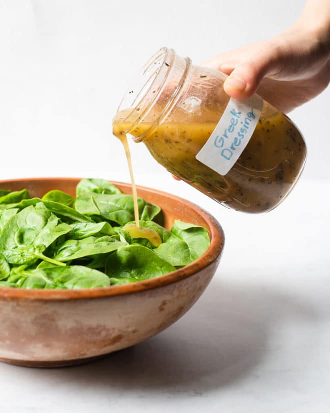 a person pouring homemade Greek dressing over a wooden bowl of spinach leaves