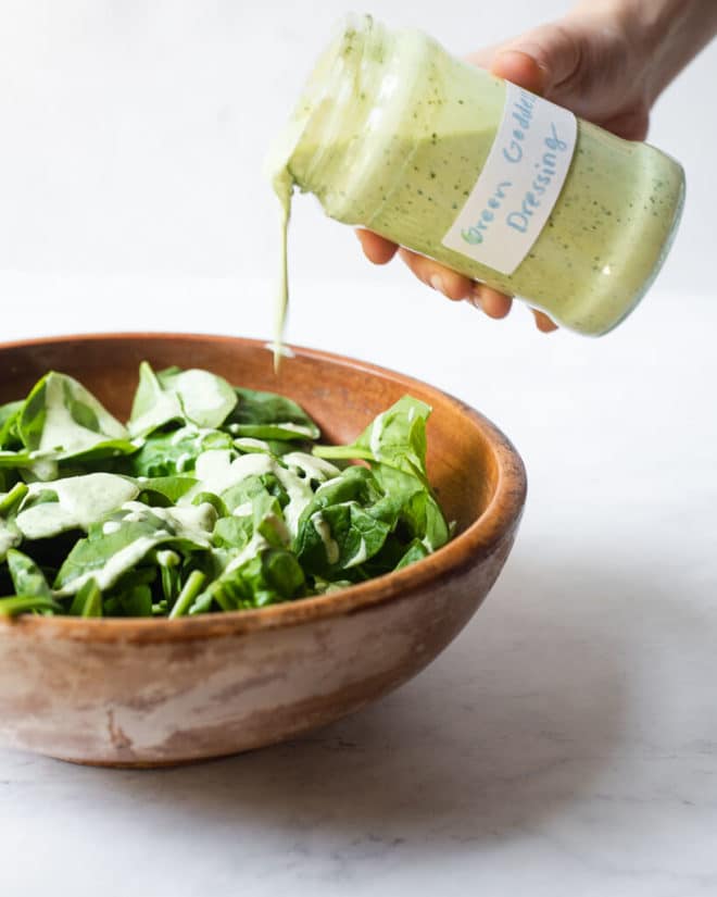 a person pouring creamy green goddess dressing over a wooden bowl of spinach leaves