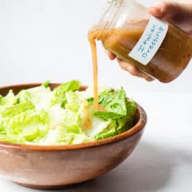a person pouring italian dressing over a bowl of chopped romaine lettuce