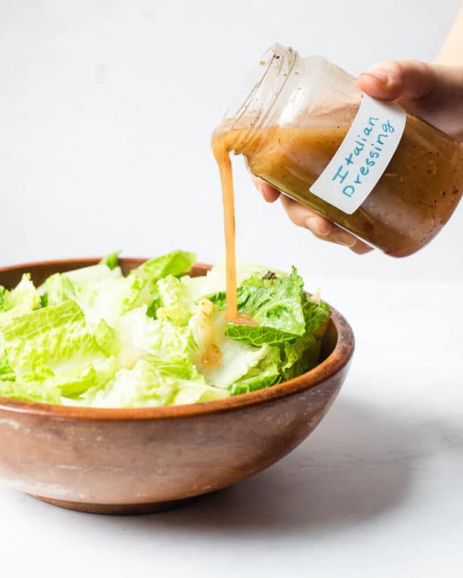 a person pouring italian dressing over a bowl of chopped romaine lettuce