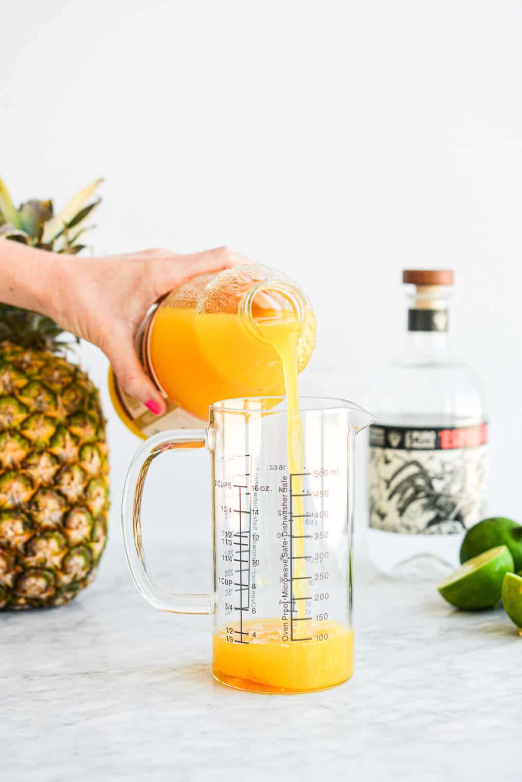 a person pouring pineapple juice into a large glass measuring cup along with the other pineapple margarita ingredients