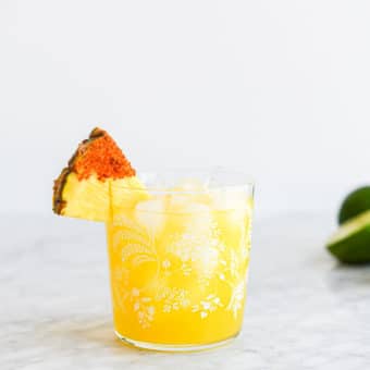 a margarita glass with a pineapple margarita on the rocks in it garnished with a pineapple wedge dipped in chili salt