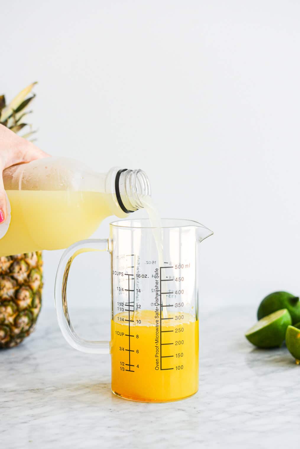 a person pouring lime juice into a large glass measuring cup along with the other pineapple margarita ingredients