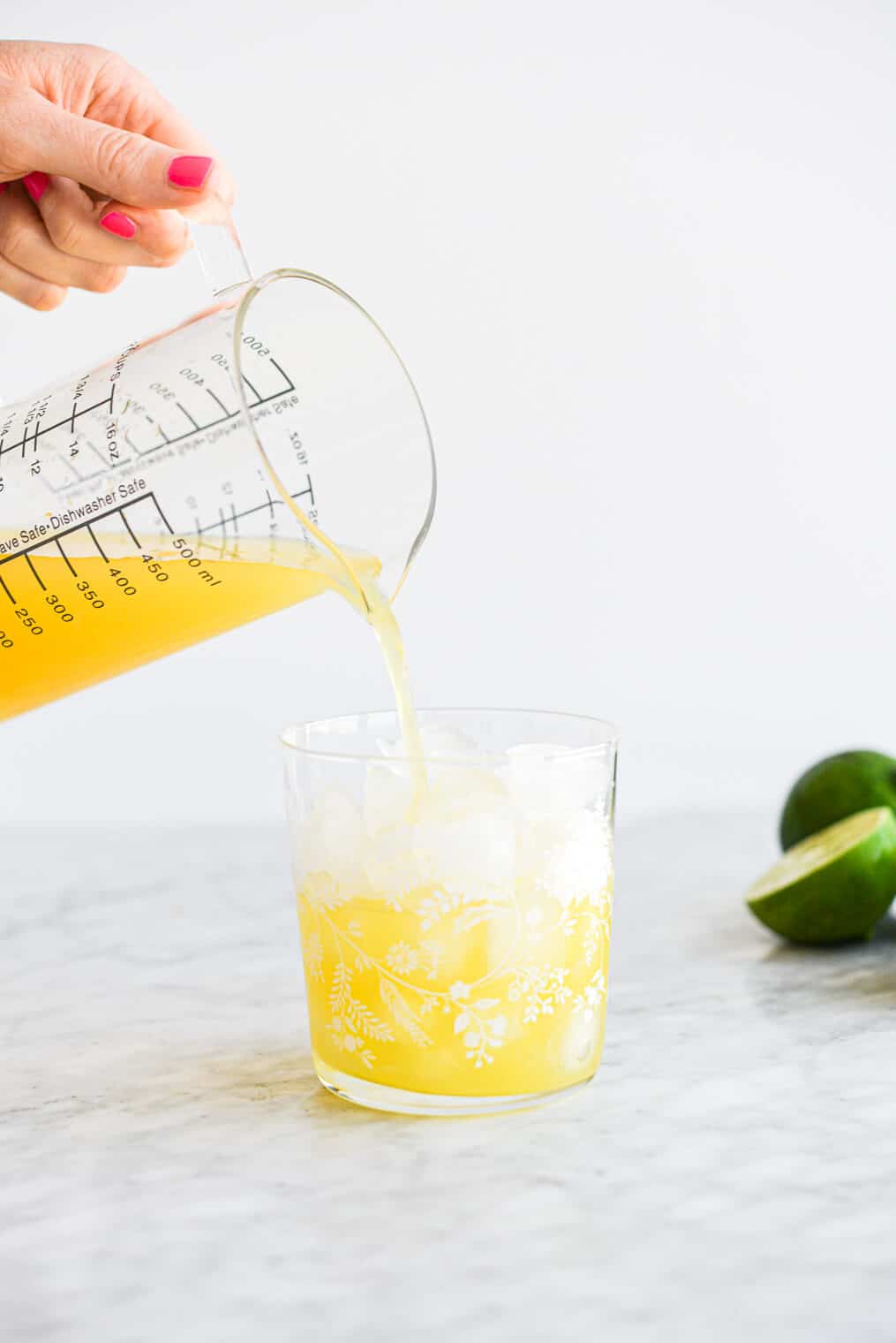 a person pouring a pineapple margarita from a large measuring cup into a margarita glass filled with ice