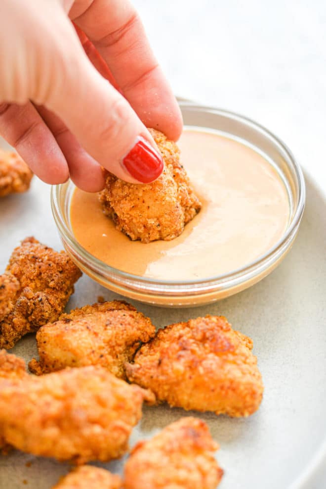 a person dipping an air fried chicken nugget into a small bowl of orange sauce