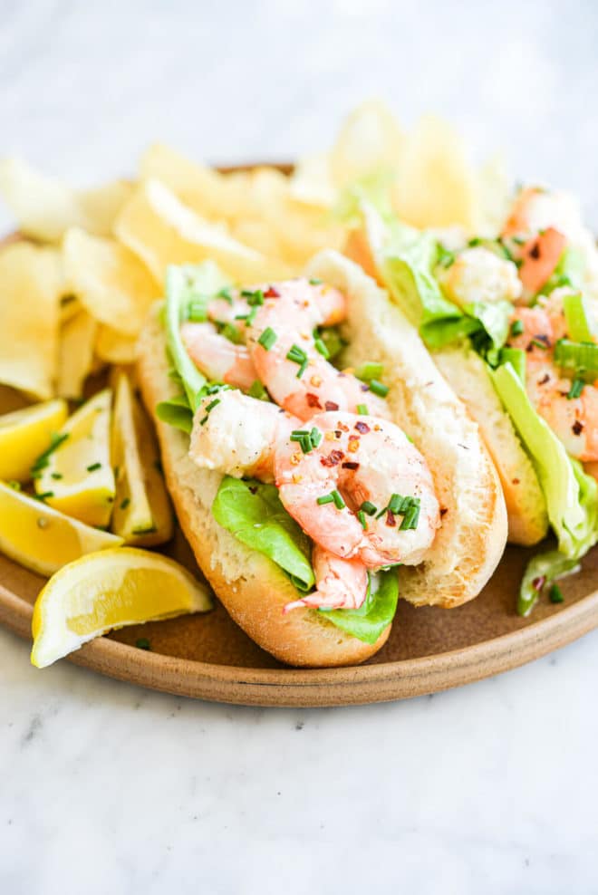 two rolls of bread filled with lettuce leaves and a boiled shrimp salad on a plate next to potato chips and lemon wedges