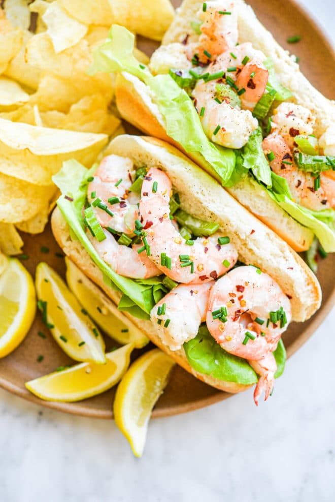 two rolls of bread filled with lettuce leaves and a boiled shrimp salad on a plate next to potato chips and lemon wedges