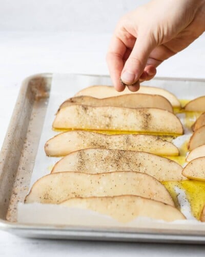 a person sprinkling ground pepper olive oil coated potato wedges on a sheet pan