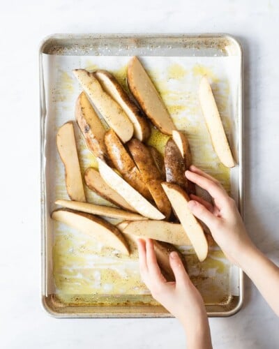 a person tossing potato wedges with olive oil, salt, and pepper on a sheet pan