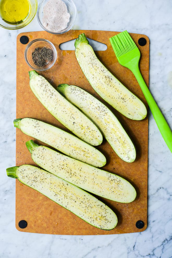 a cutting board of halved raw zucchini sitting next to a basting brush and three small bowls of salt, pepper, and olive oil