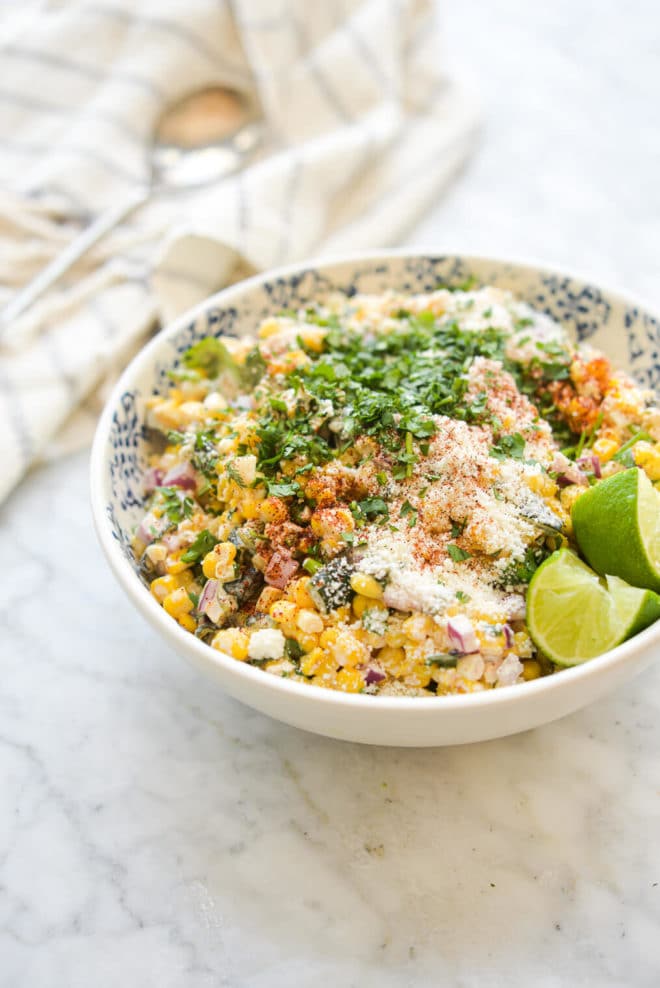 a bowl of mexican street corn salad garnished with cotija cheese, cilantro, lime wedges, and tajin