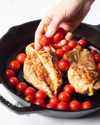 a person sprinkling cherry tomatoes around two seared chicken breasts in a cast iron skillet