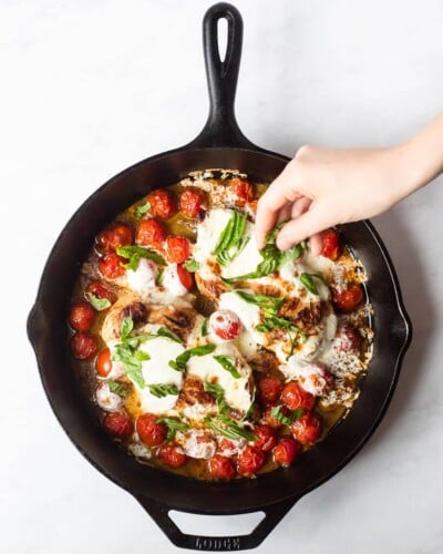 a person sprinkling fresh basil over top a cooked caprese chicken skillet with chicken breasts, bursted cherry tomatoes, and melted mozzarella cheese