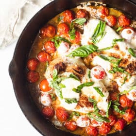 a cooked caprese chicken skillet with chicken breasts, bursted cherry tomatoes, melted mozzarella cheese, and fresh basil