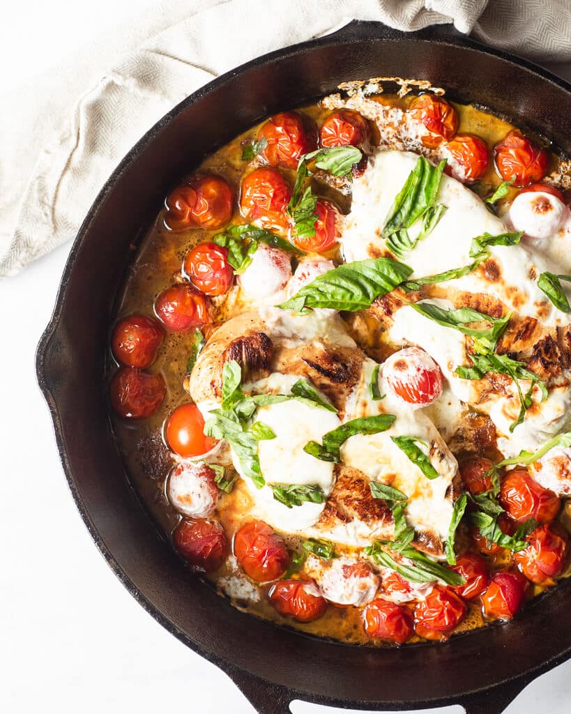 a cooked caprese chicken skillet with chicken breasts, bursted cherry tomatoes, melted mozzarella cheese, and fresh basil