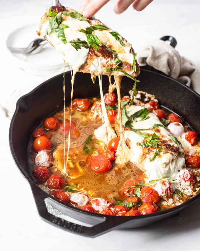 a person pulling a mozzarella covered seared chicken breast out of a caprese chicken skillet to serve