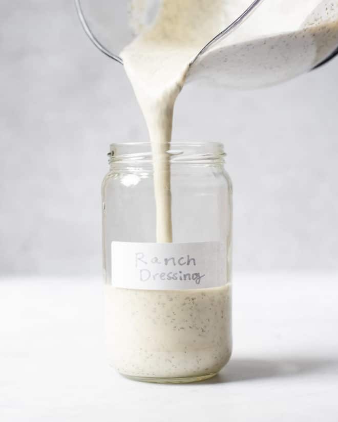 a person pouring homemade ranch dressing from a large, spouted measuring cup into a labeled jar