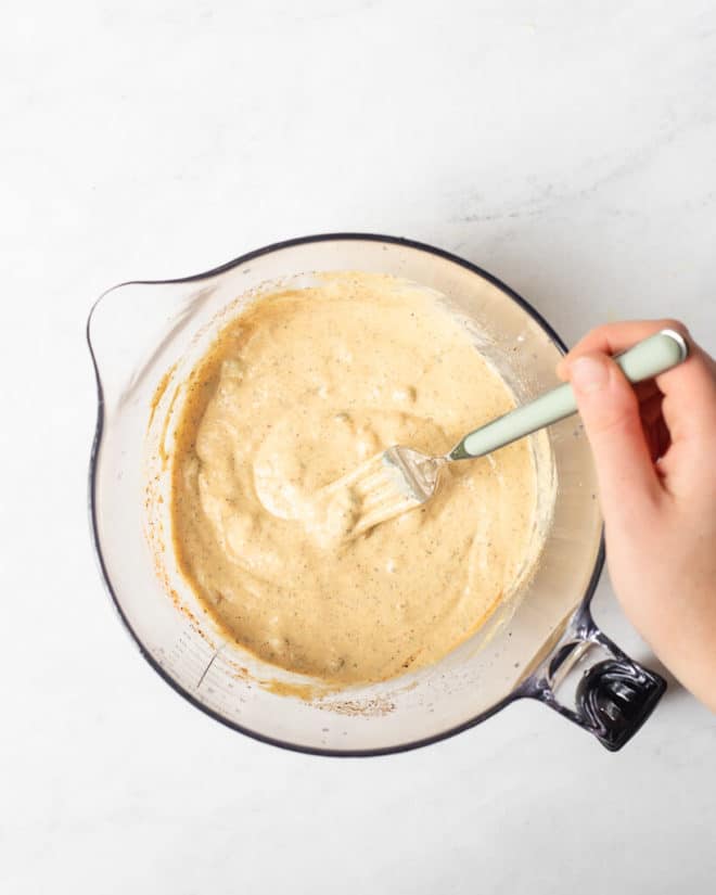 a person using a fork to whisk together a homemade remoulade sauce in a large, spouted, glass mixing bowl