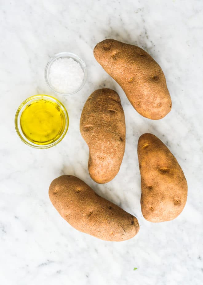 4 russet potatoes laying next to a bowl of olive oil and a bowl of coarse sea salt