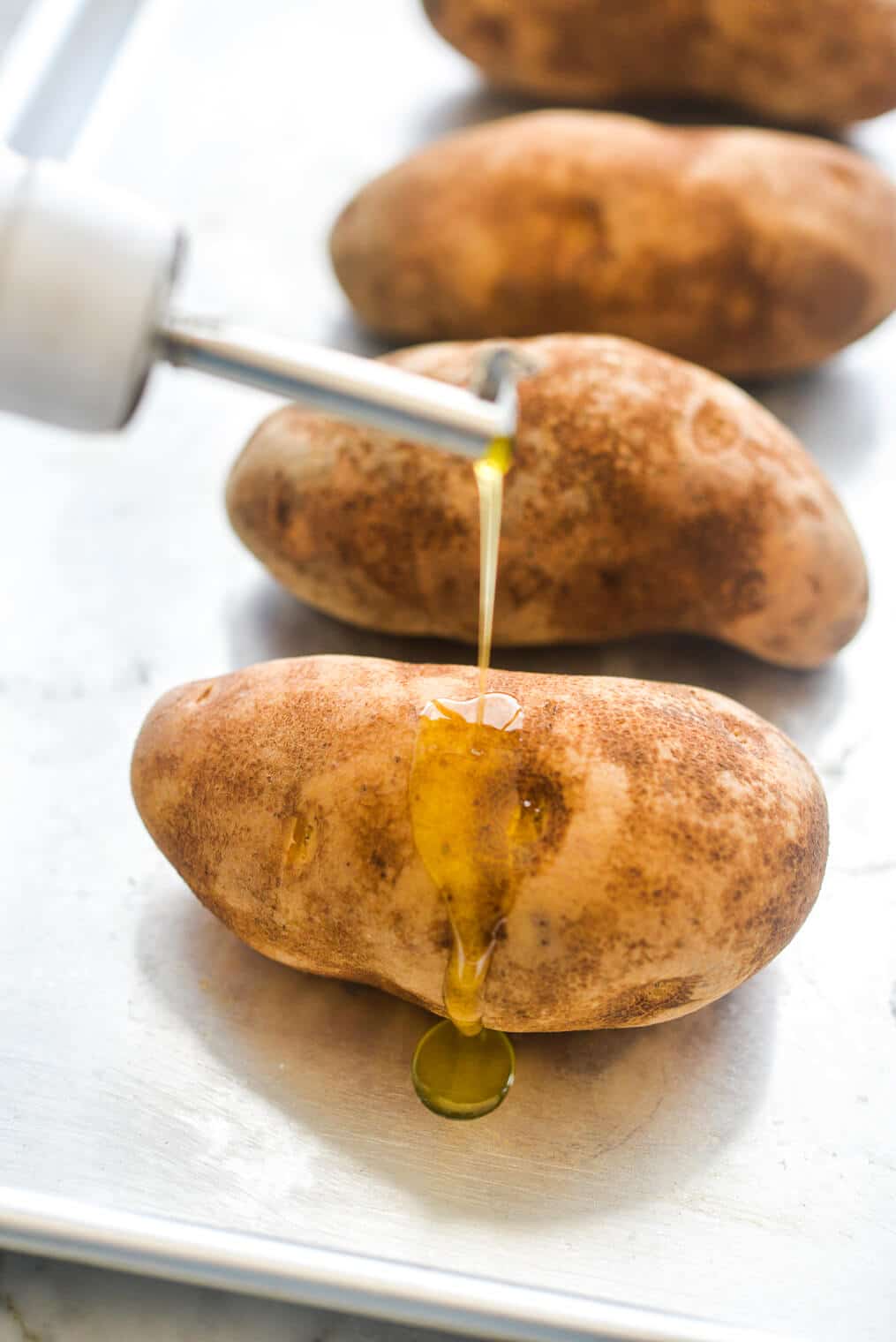 4 raw russet potatoes being drizzled with olive oil on a metal sheet pan