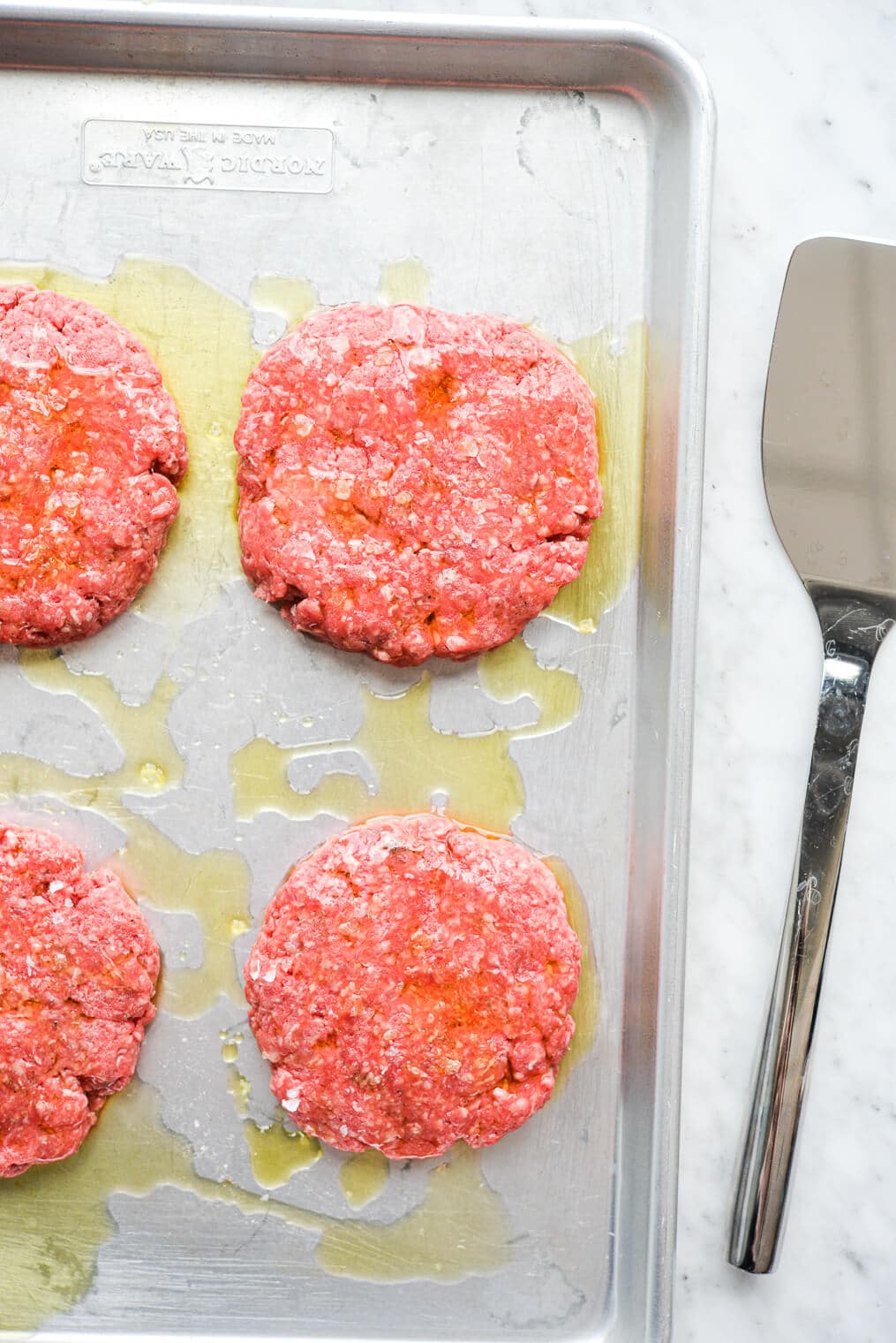 raw beef patties drizzled with olive oil sitting on a metal sheet pan
