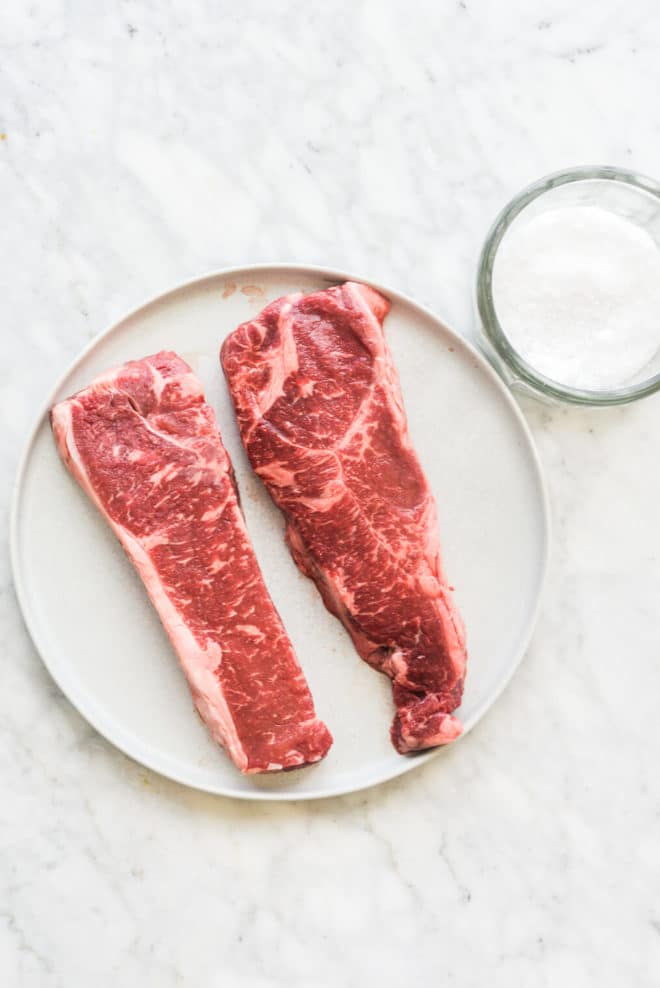 two raw steaks sitting on a plate next to a bowl of coarse sea salt