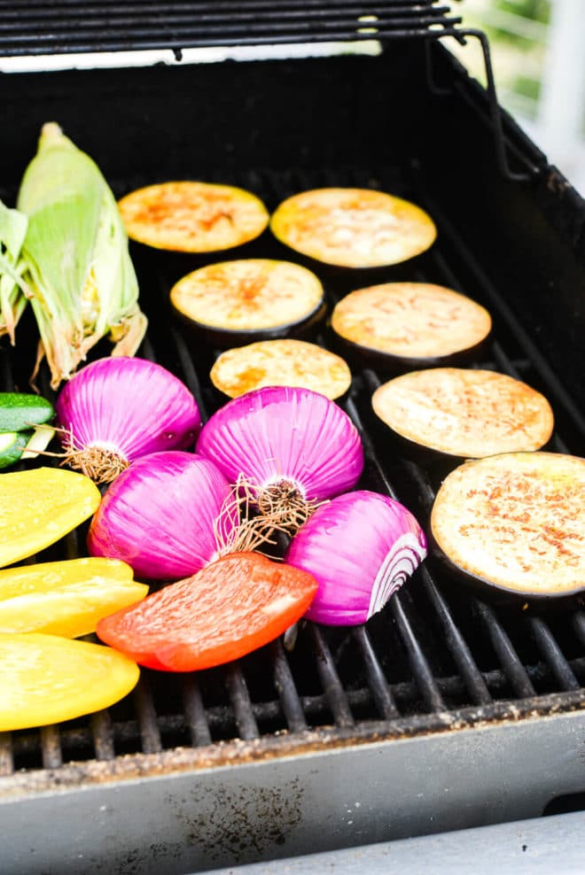 sliced eggplant, sliced bell pepper, halved onions, and corn on the cob all on the grill grates of a gas grill