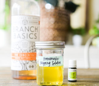 a bottle of Branch Basics Concentrate, essential oil, and a mason jar of mopping solution sitting on a wooden kitchen table