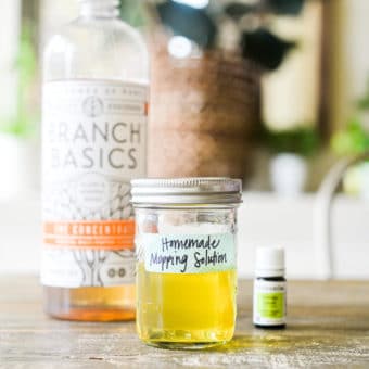 a bottle of Branch Basics Concentrate, essential oil, and a mason jar of mopping solution sitting on a wooden kitchen table