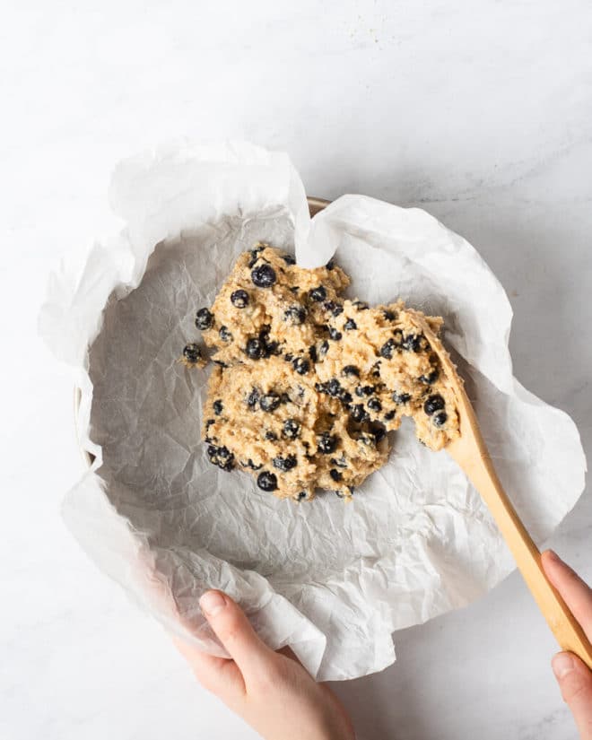 a person spooning blueberry scone batter into a parchment paper lined circular baking dish