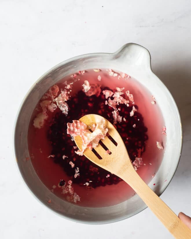 pomegranate seeds in the bottom of a bowl of water with the pith of the pomegranate floating to the top and being scooped out by a wooden spoon