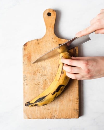 a person cutting the tip off of a plantain on a wooden cutting board using a large knife