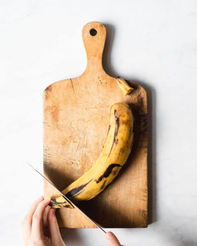 a person cutting the tip off of a plantain on a wooden cutting board using a large knife