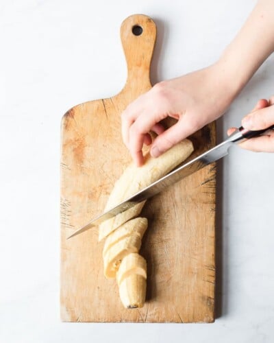 a person slicing a plantain on a wooden cutting board using a knife