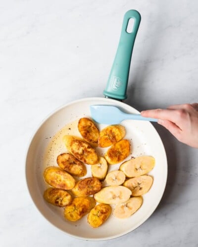someone using a small blue spatula to pan fry sliced plantains