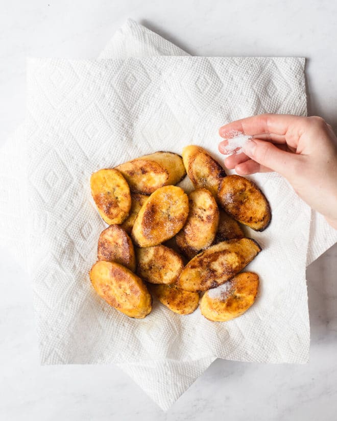 a person sprinkling salt onto sliced, pan fried plantains on a paper towel lined plate