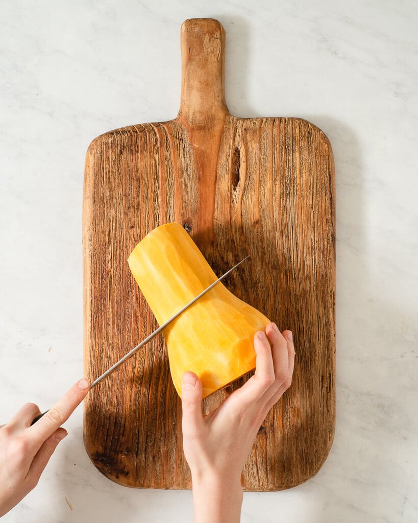 a person cutting a peeled butternut squash on a wooden cutting board using a large knife