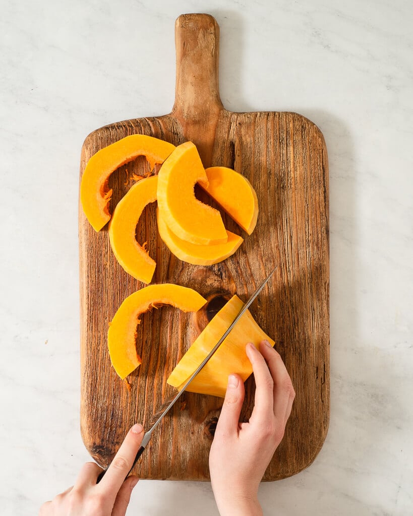 a person cubing butternut squash on a wooden cutting board using a large knife