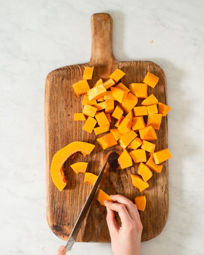 a person cubing butternut squash on a wooden cutting board using a large knife