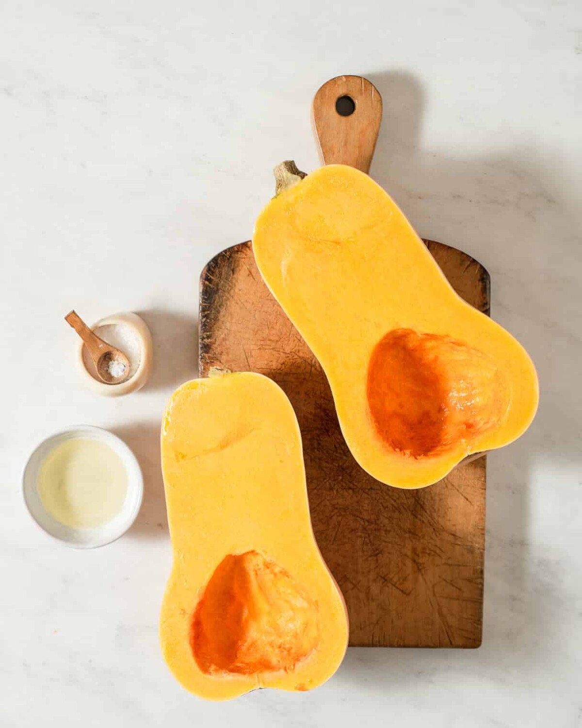 two butternut squash halves on a wooden cutting board next to a small bowl of salt and a small bowl of oil