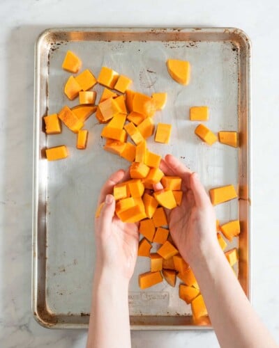 a person tossing cubed butternut squash with olive oil onto a sheet pan