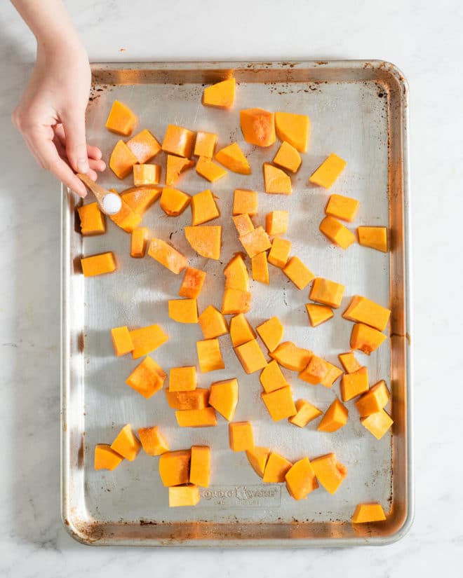 cubed, raw butternut squash on a sheet pan being sprinkled with sea salt