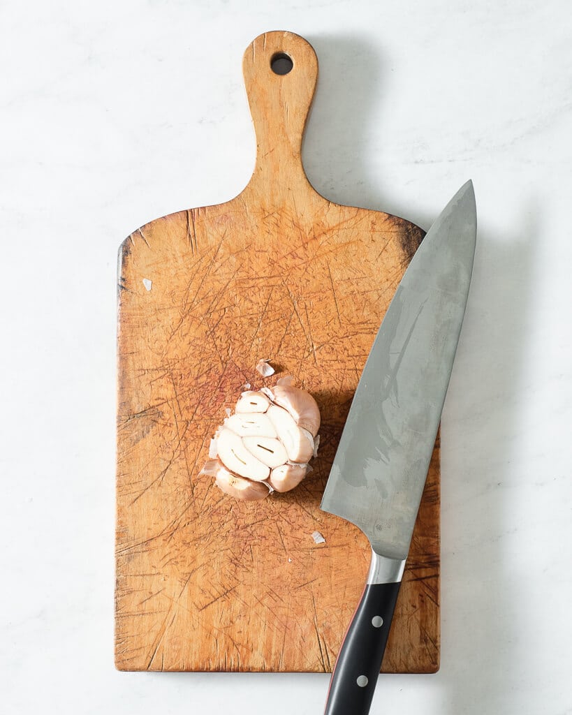 a bulb of garlic with the top cut off sitting on a wooden cutting board next to a knife