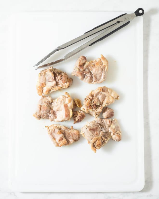 chicken thighs on a cutting board next to stainless steel tongs